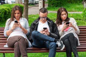 three people on bench looking down at cell phone with tech neck